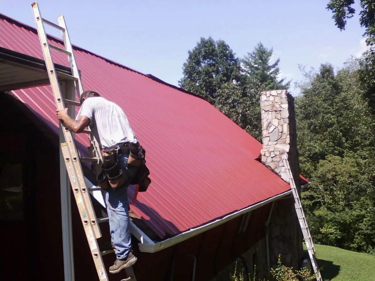 Roofer on a ladder beside a red metal roof and stone chimney — Soffit & Fascia Repair by Roofing Brothers in Westfir